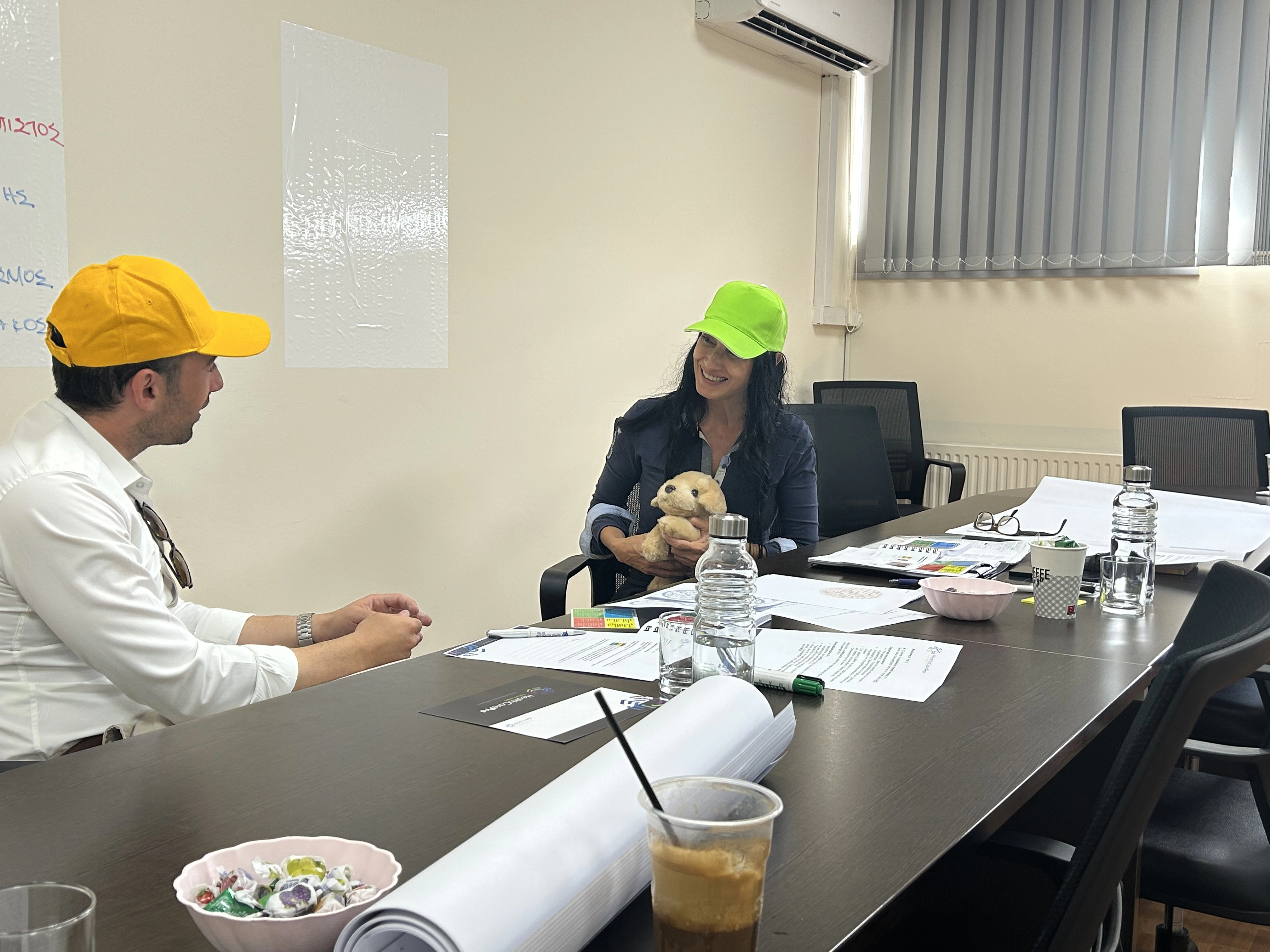 A man and a woman seated around a desk, discussing professionally, with the woman holding a teddy bear as part of a workshop exercise.
